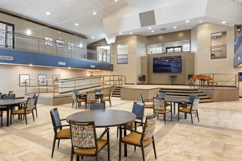 Large open activity hall with round tables and chairs, a stage area and a balcony above