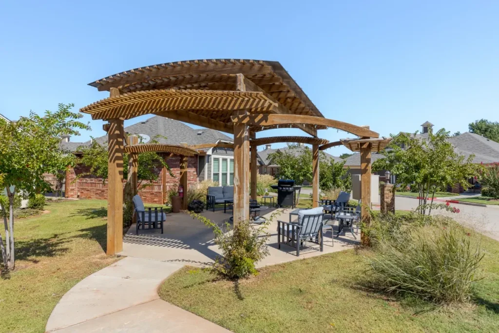 Wooden pergola over a patio with tables and chairs, surrounded by lawn and shrubs
