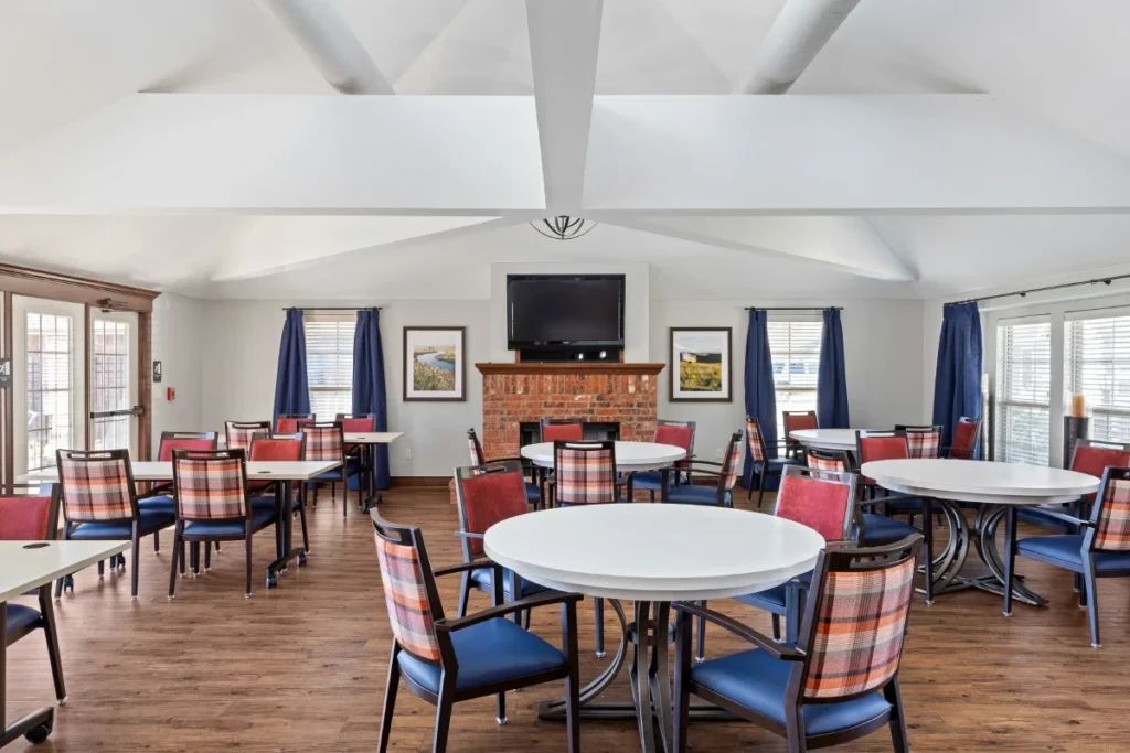 Dining room with round tables, plaid chairs, a fireplace with a TV and blue curtains at tall windows