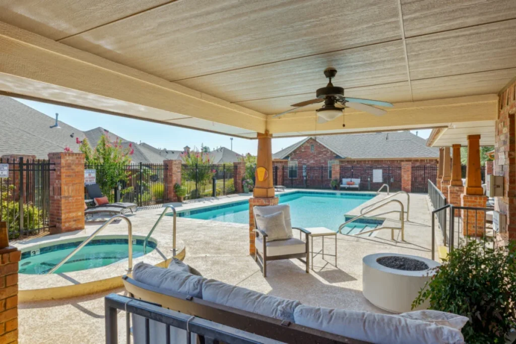 Covered poolside patio with a ceiling fan, seating and a fire pit, with a swimming pool and spa beyond