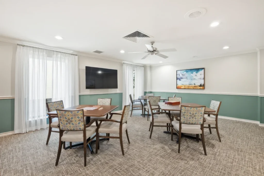 Activity room with tables and chairs, a wall-mounted TV and sheer curtains at a window