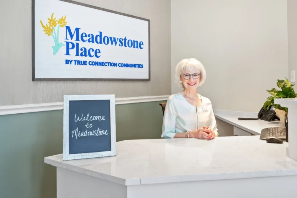 Smiling receptionist at a white front desk under a wall-mounted sign, with a small welcome chalkboard
