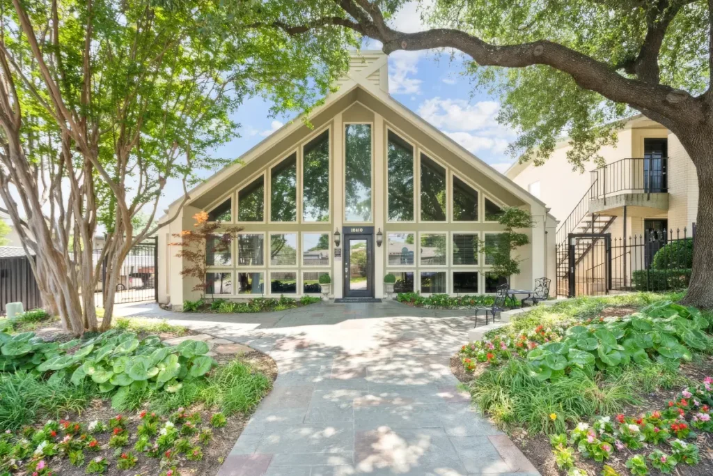 Glass-fronted building with a tall peaked roof, framed by trees and flower beds along a walkway