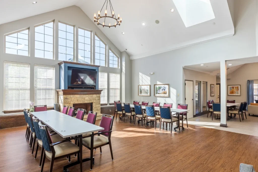 Mon Abri dining and activity room with long tables, chairs, a stone fireplace with a TV and tall windows under a vaulted ceiling