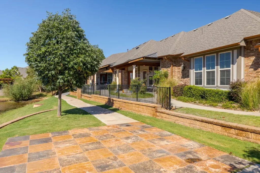 Stone patio and lawn beside a row of single-story brick homes, with a tree in the foreground