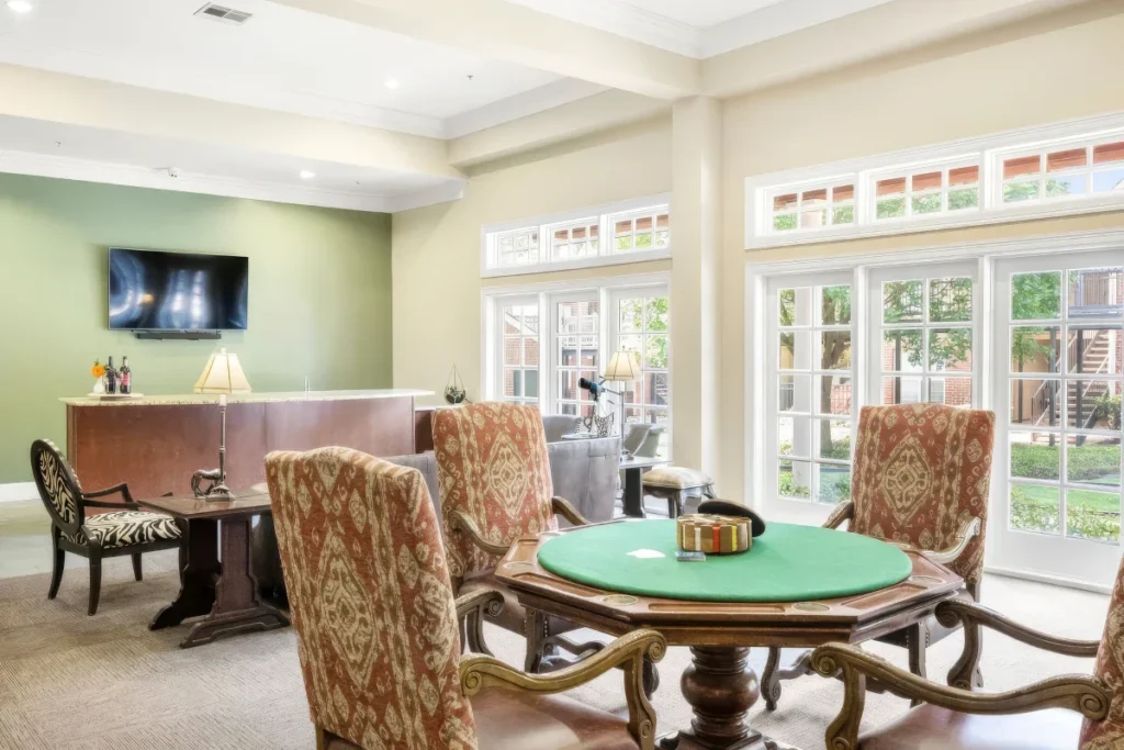 Lounge with a round green-topped game table, upholstered chairs and a wall-mounted TV, near large glass doors