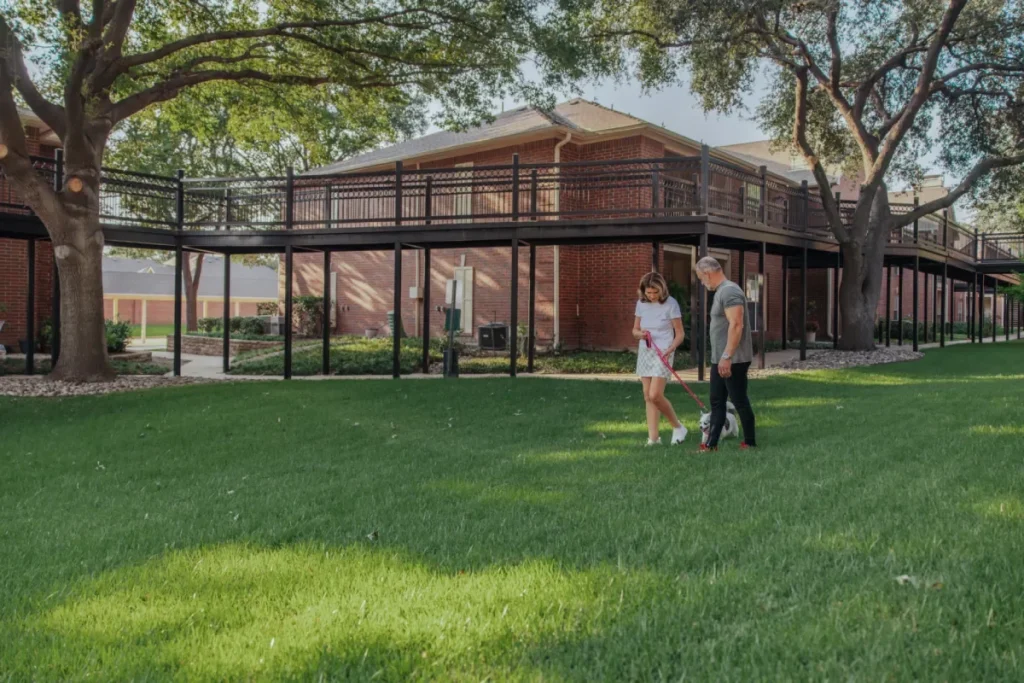 Two people walking across a lawn shaded by large trees, with brick apartment buildings and covered walkways behind