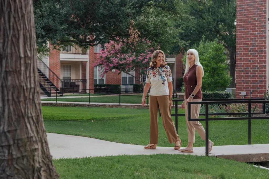 Two women strolling on a walkway beside a lawn and flowering trees, with brick buildings behind