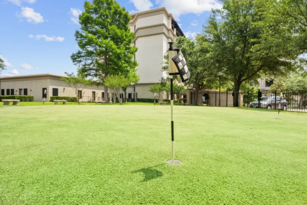 Grassy putting green with a flagstick, surrounded by trees, with a building beyond