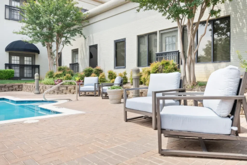 Poolside patio with white lounge chairs and a small pool, in front of a building with landscaped beds