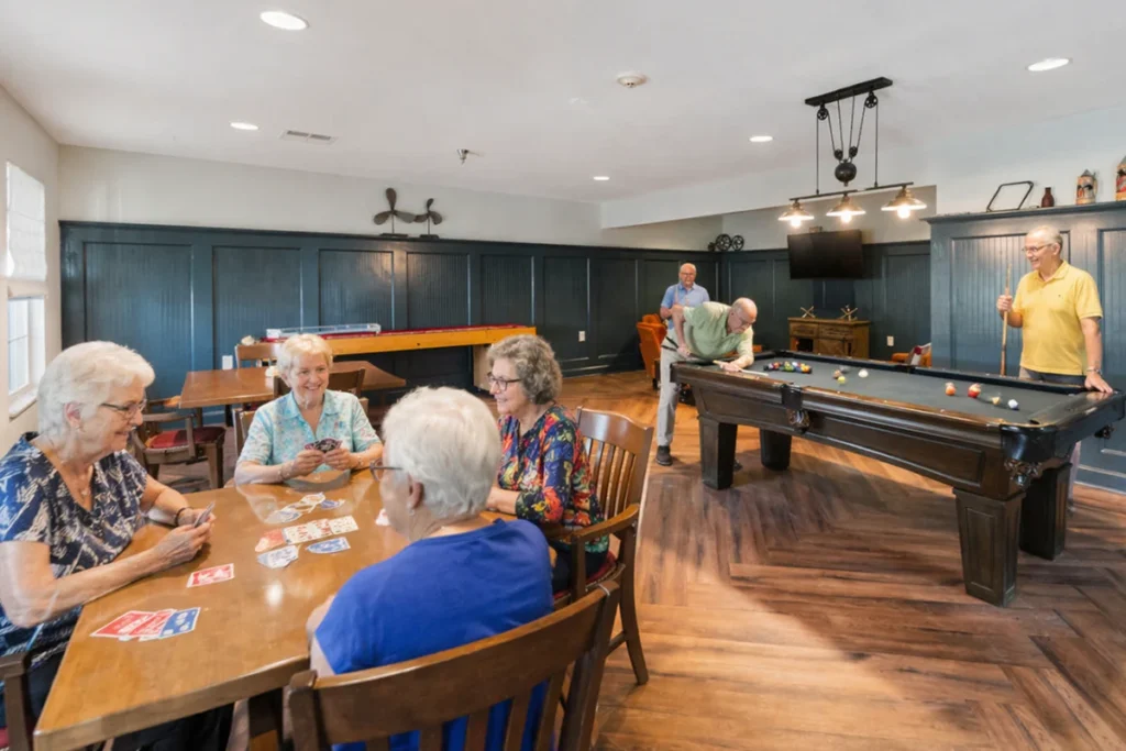 Older adults playing cards at a table in a game room with a pool table behind them