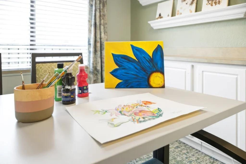 Art table with a painted blue flower, a watercolor sheet, paint bottles and a cup of brushes