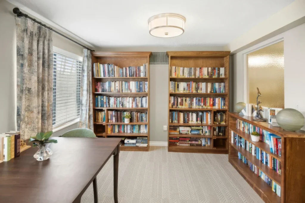 Library room with wooden bookcases full of books, a table and window curtains