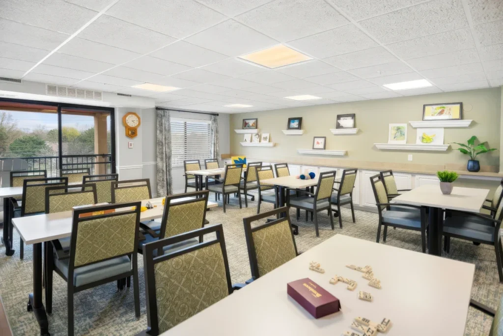 Activity room with tables and chairs, a small game box on a table in the foreground, and windows overlooking trees