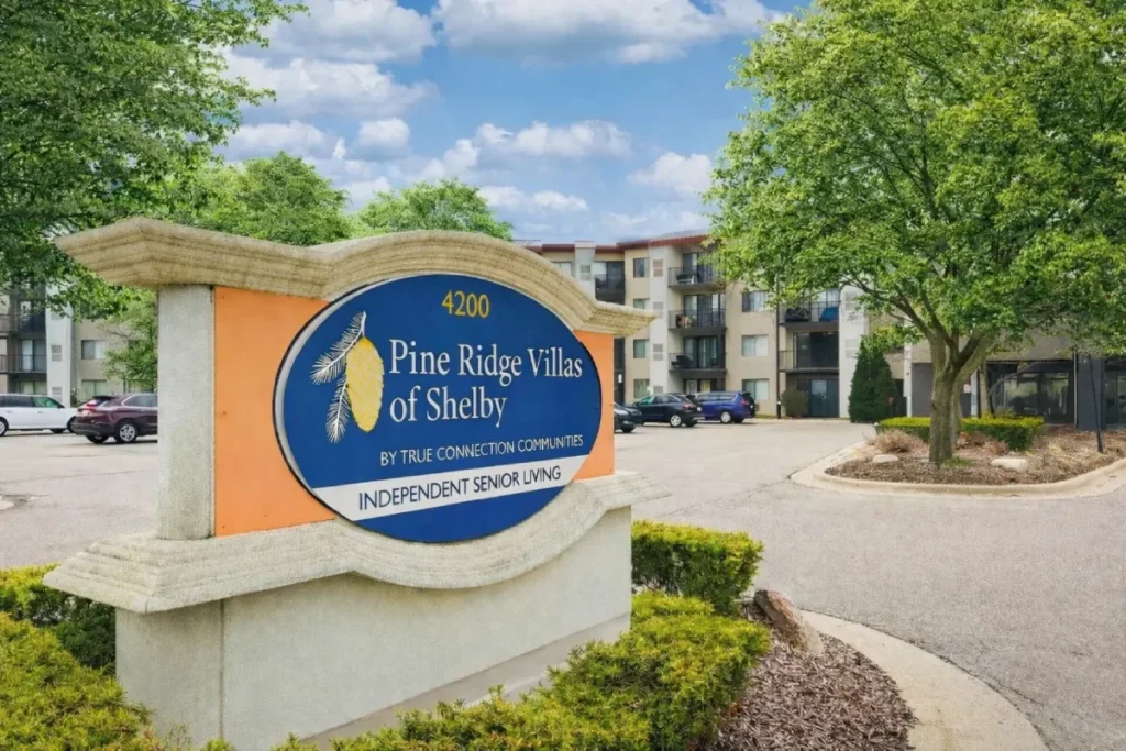 Entrance monument sign with a blue and orange emblem, surrounded by landscaping, with an apartment building and trees behind