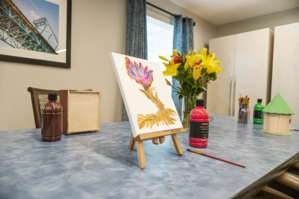 Small easel with a painted flower beside a vase of yellow flowers and paint bottles on a craft table