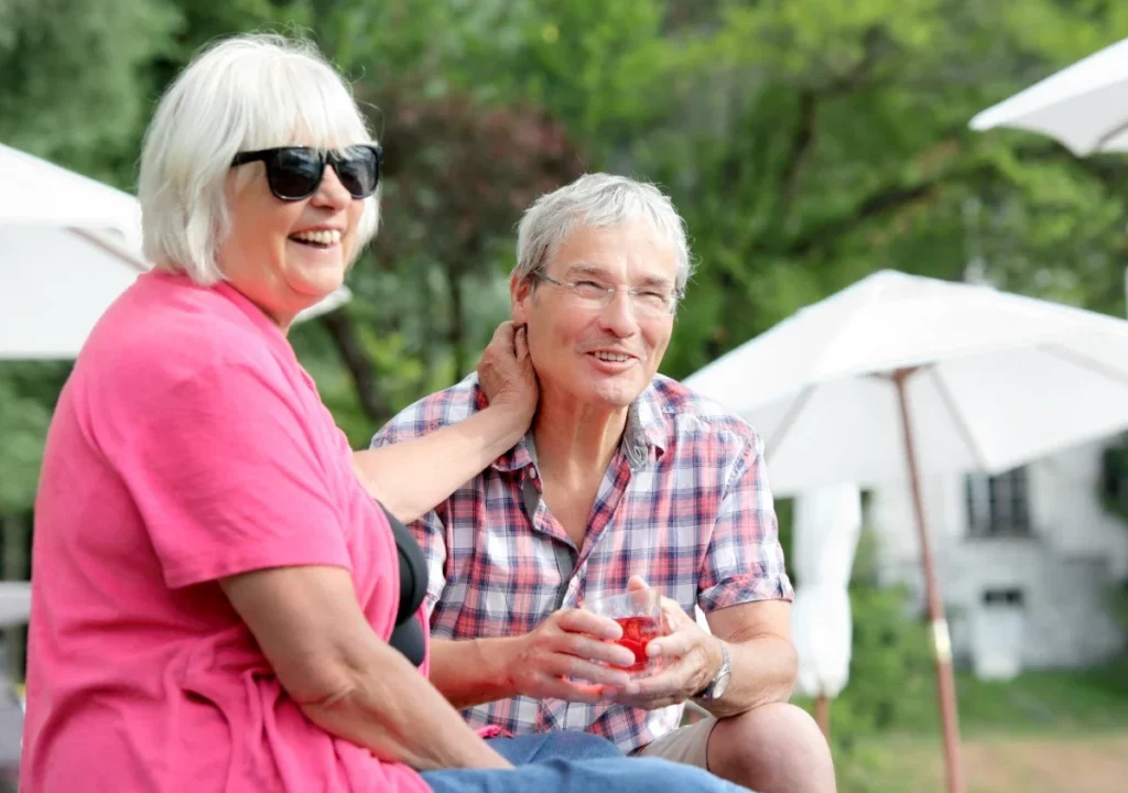 Two older adults laughing together outdoors with drinks under patio umbrellas