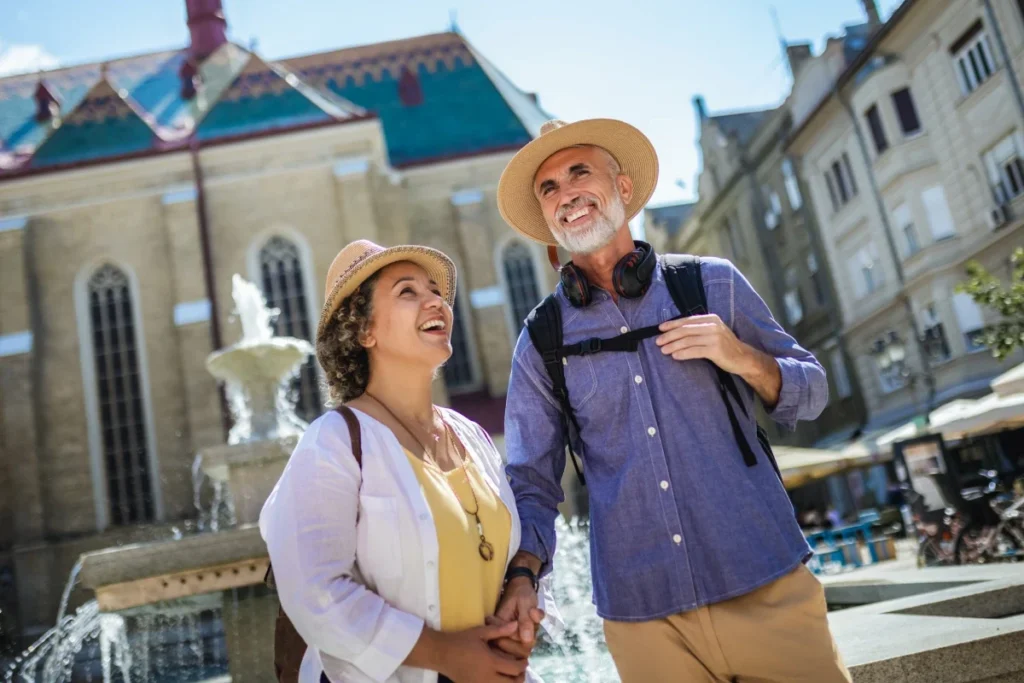 Smiling older couple in sun hats and backpacks sightseeing beside a fountain in front of an ornate historic building