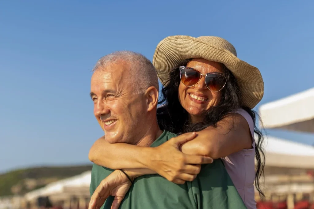 Older woman in a straw hat and sunglasses hugging a smiling older man from behind, with beach loungers in the background