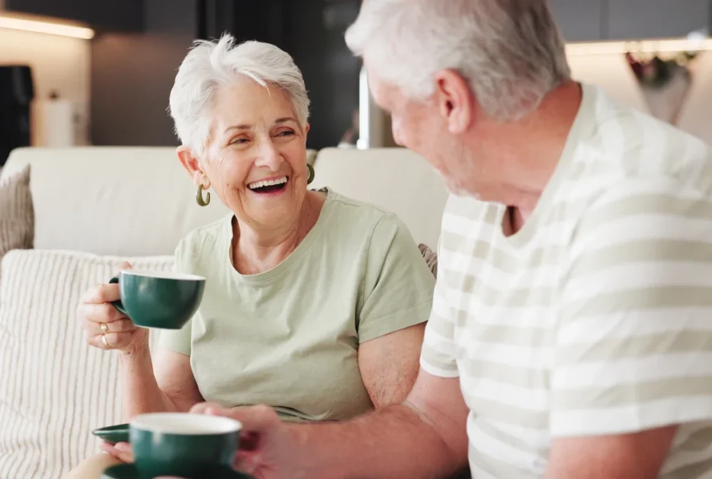 Smiling older woman and man laughing while holding green mugs on a sofa