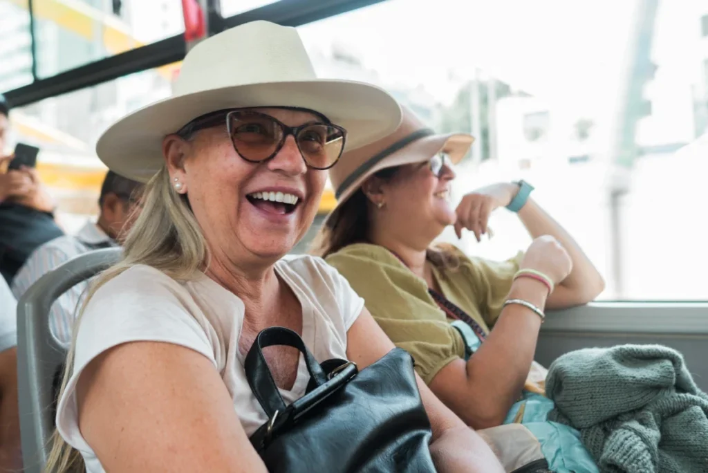 Two smiling older women in sun hats seated on a bus, one laughing toward the camera