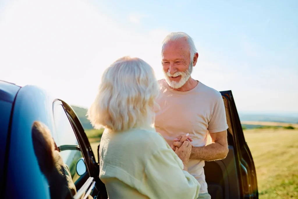 Older couple standing beside a car with an open door, looking at each other with a field behind them