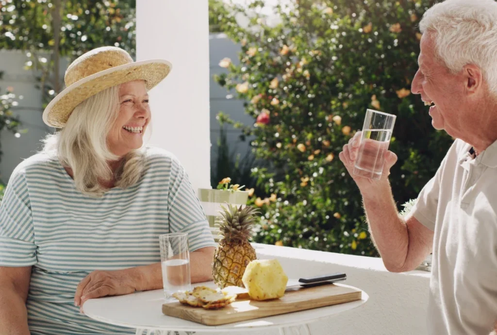 Smiling older woman in a straw hat and an older man raising a glass, at a table with a pineapple and fruit board