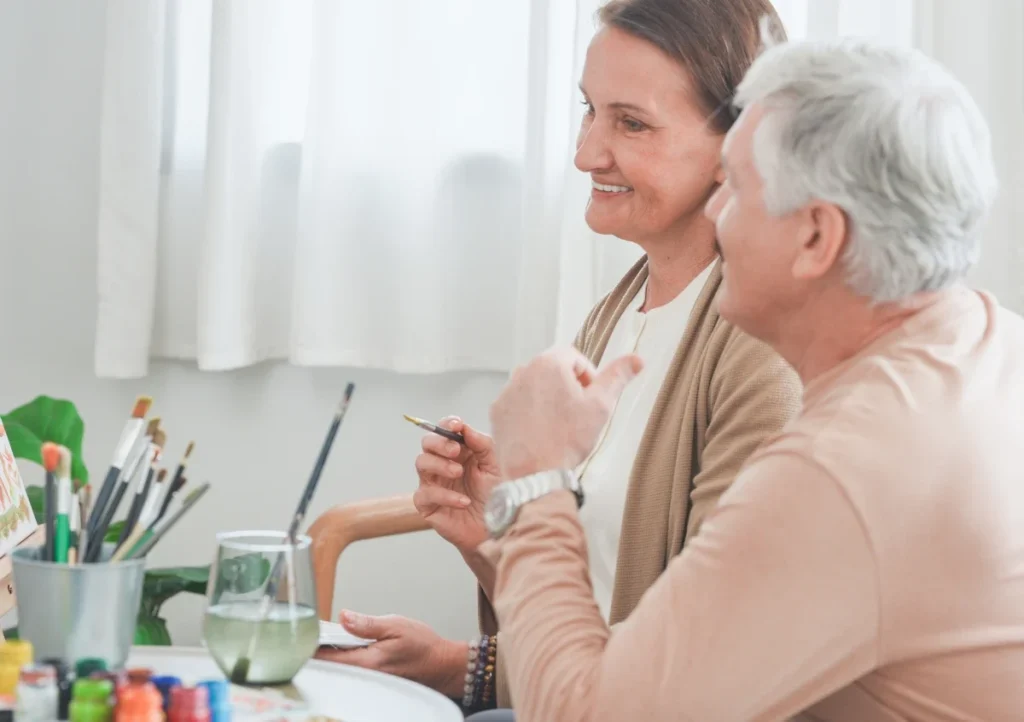 Two older adults painting together at a table with brushes and jars
