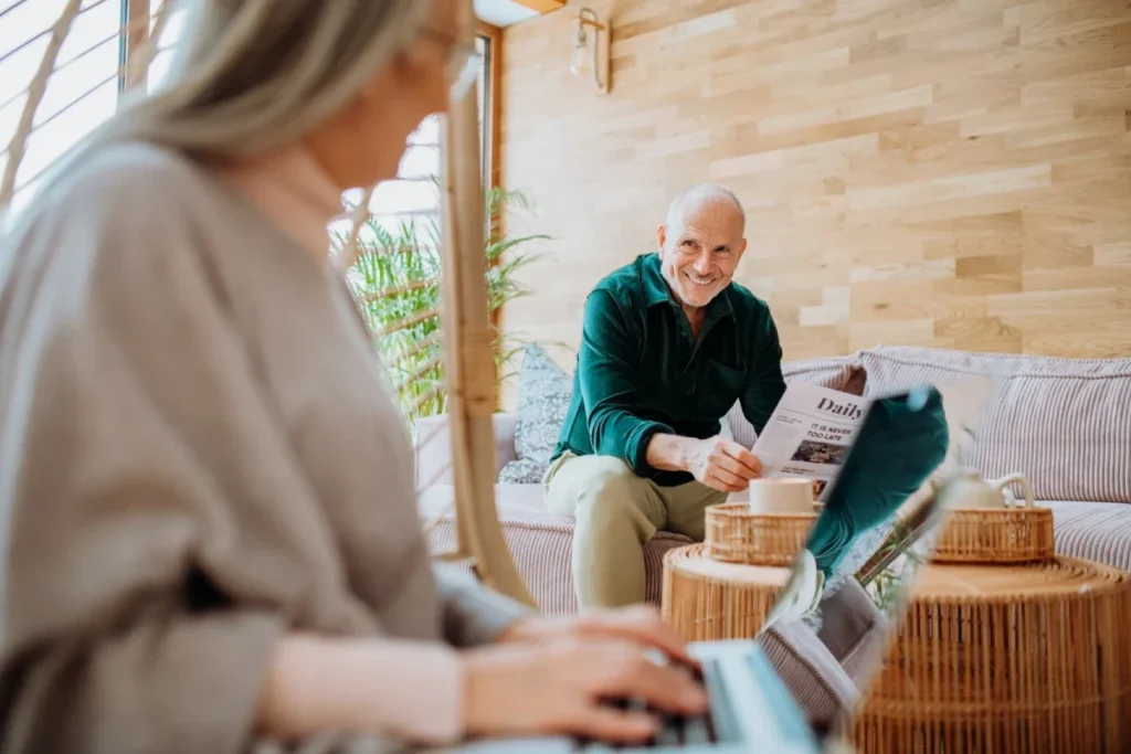 Woman typing on a laptop in the foreground, with a man reading a newspaper on a sofa behind her