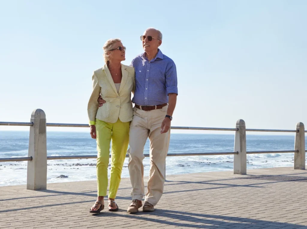 Older couple in light summer clothes standing arm in arm on a seaside promenade with the sea behind them