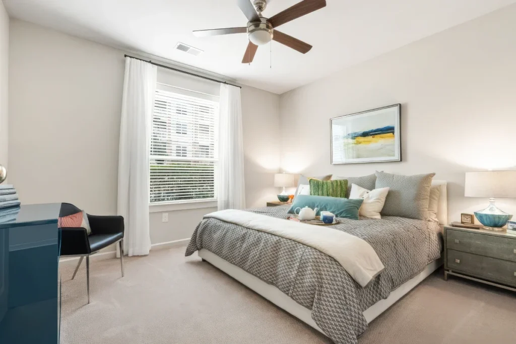 Bedroom with a gray bed, striped pillows and a ceiling fan, beside a window with white curtains