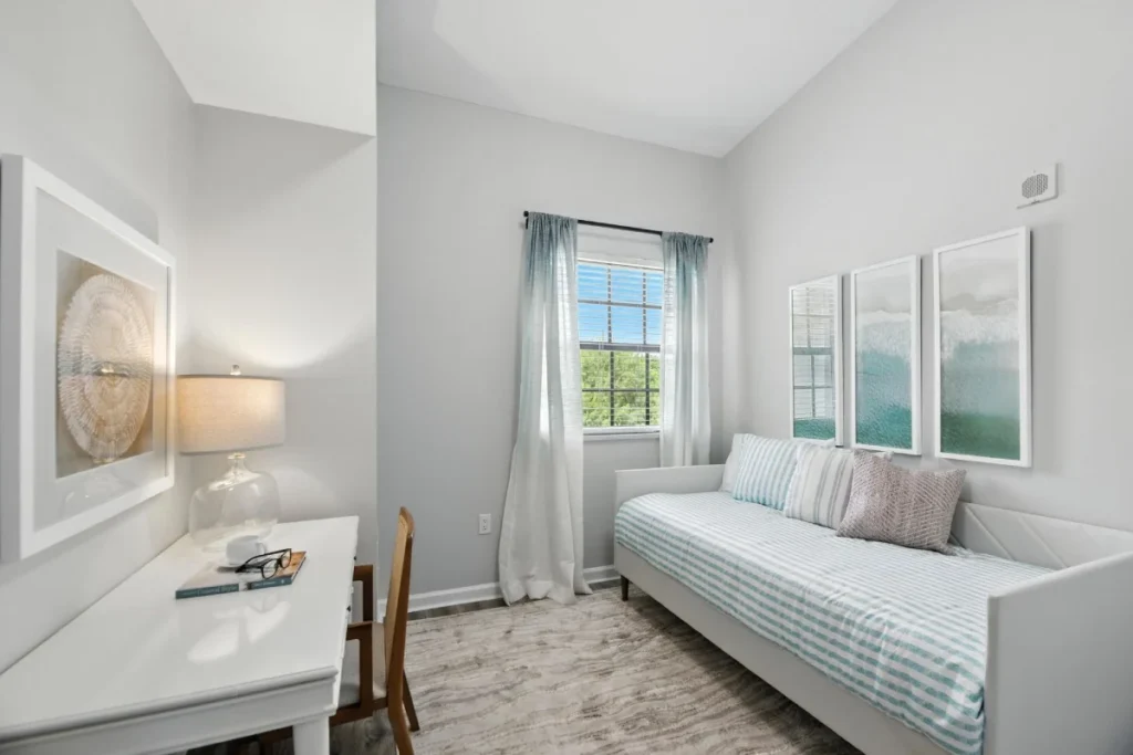 Small room with a daybed, a white desk and a lamp, with framed coastal-style art on the walls