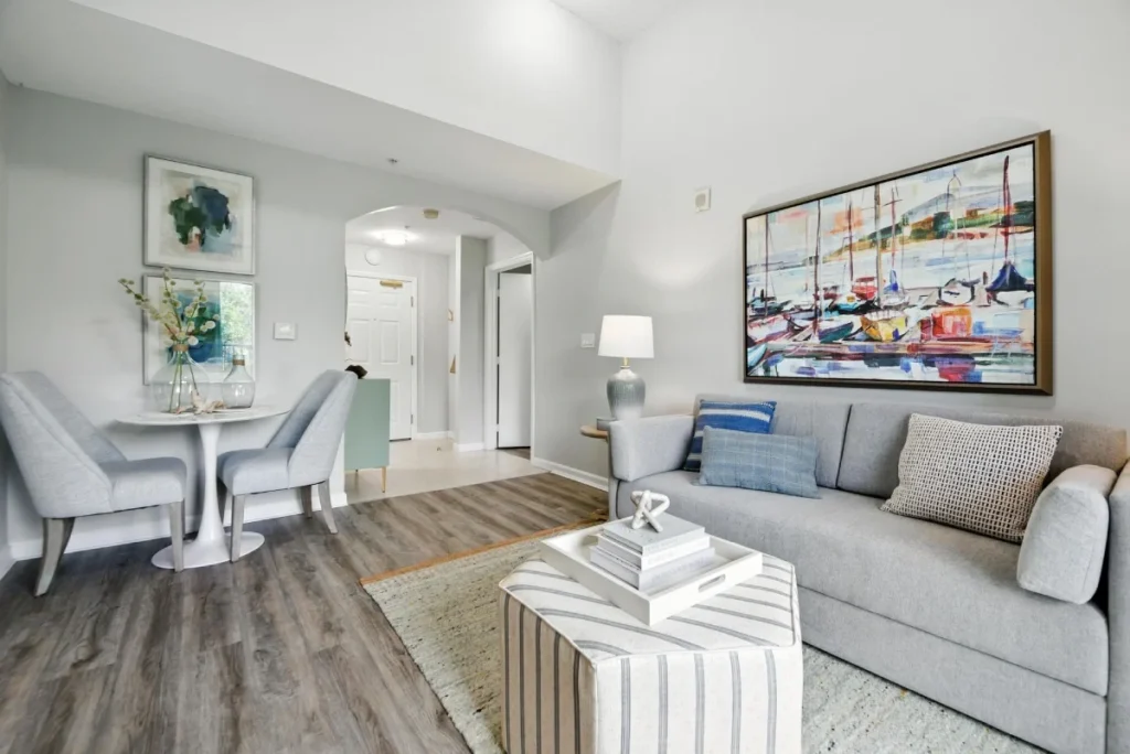 Apartment living area with a gray sofa, a colorful sailboat painting and a small dining table beyond