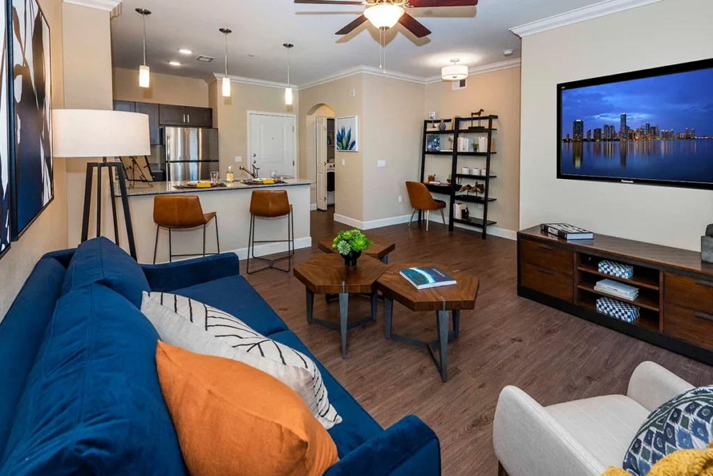 Living room with a blue sofa and orange pillows, a wall-mounted TV and an open kitchen behind
