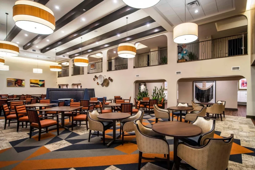 Large dining hall with round tables, upholstered chairs and pendant lights under a beamed ceiling