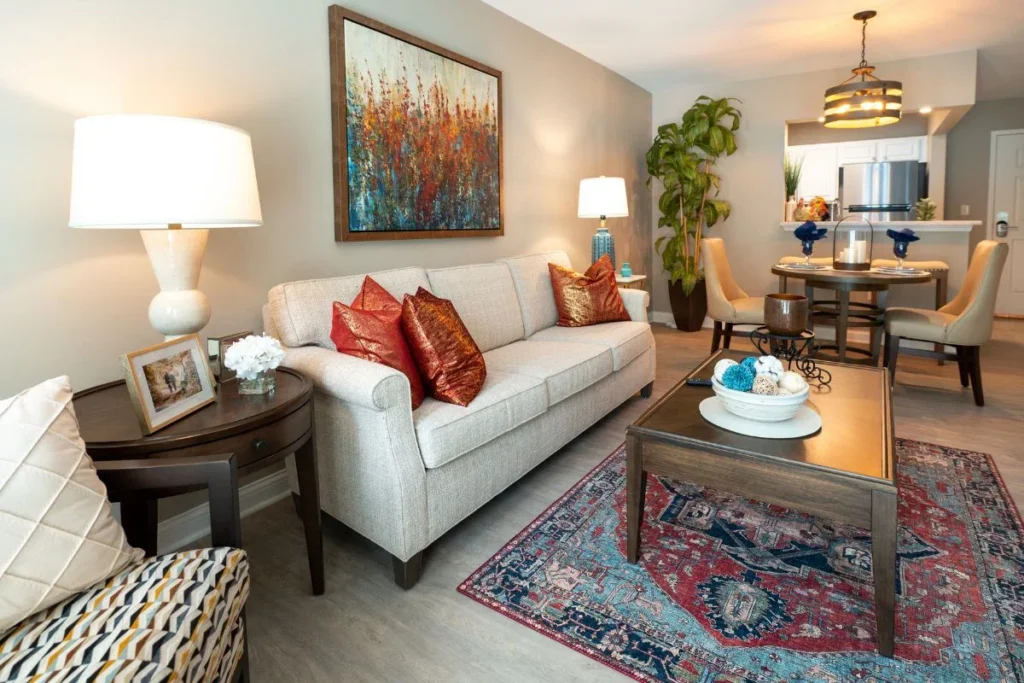 Apartment living room with a white sofa, red accent pillows and a patterned rug, with a dining table beyond