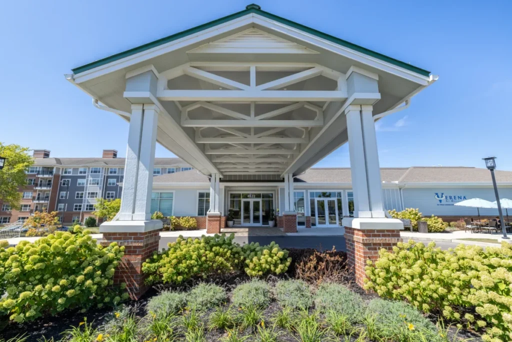 Covered entrance with white columns and brick bases at the front of a modern building, framed by flowering shrubs