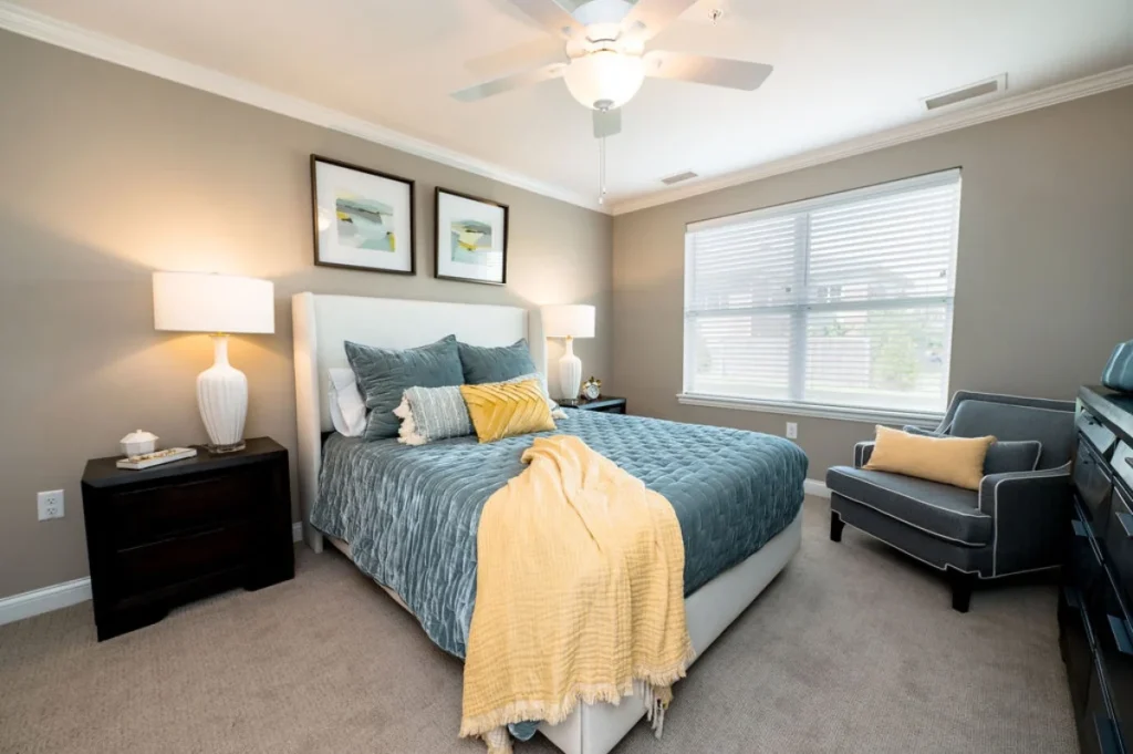 Bedroom with a made bed in teal bedding and a yellow throw, nightstands with lamps and a ceiling fan