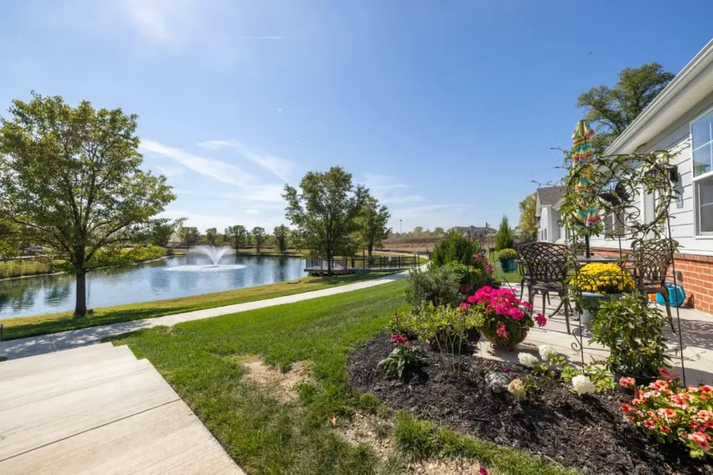 Landscaped patio with tables and flowering garden beds beside a pond and lawn