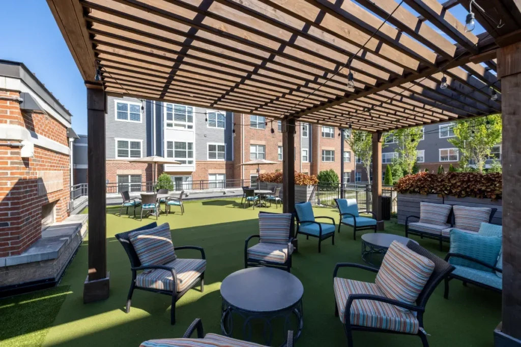 Outdoor lounge under a wooden pergola with seating on artificial turf, with brick apartment buildings behind
