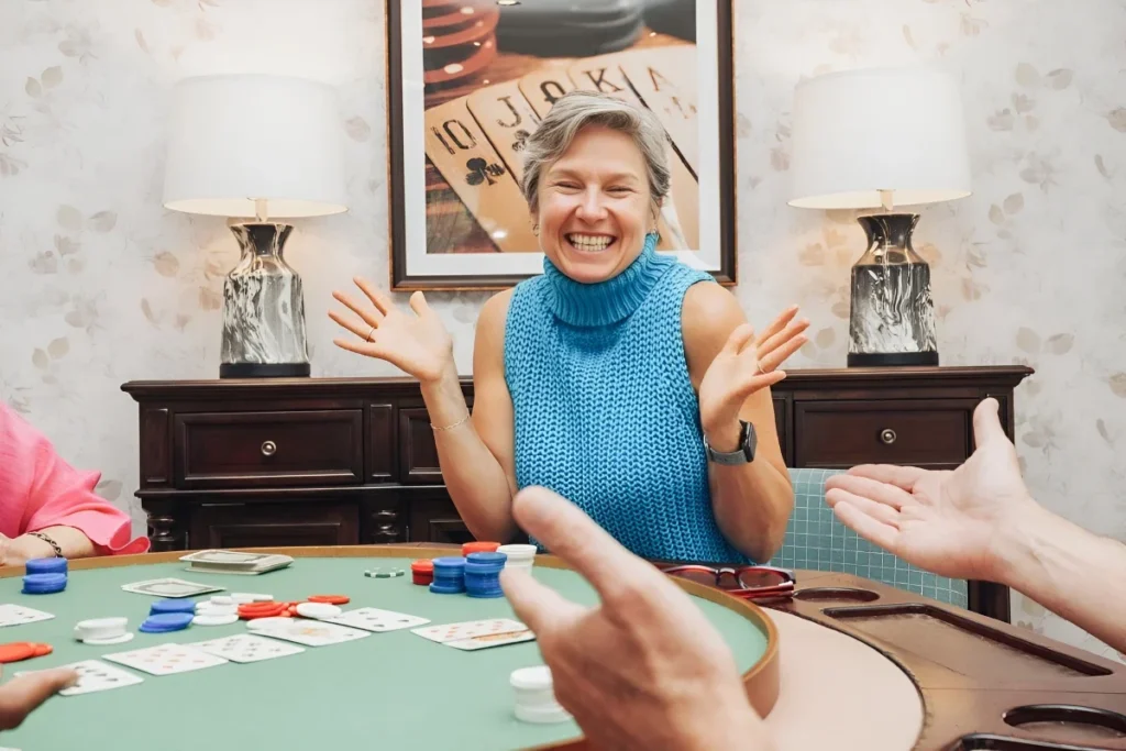 Smiling older woman in a teal top gesturing at a card table with poker chips and cards