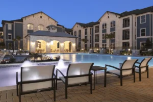 Apartment community exterior at dusk with a lit pool area and lounge chairs