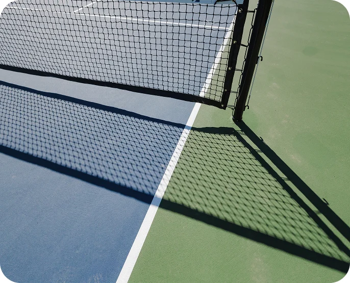 A close-up view of a tennis court net casting a shadow on the blue and green surface, with white boundary lines visible under bright sunlight—perfect for active senior living and promoting independence.