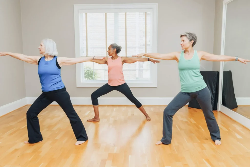 Three older women practicing yoga warrior pose in a bright studio