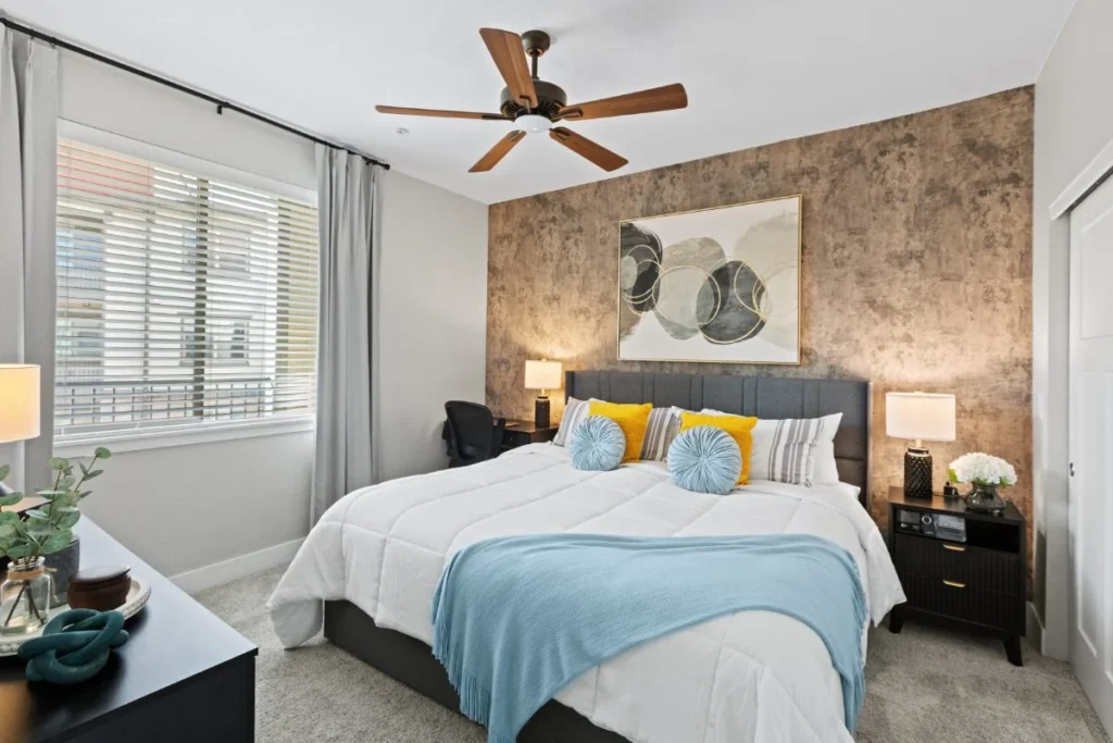 Bedroom with a ceiling fan, a bed with blue and yellow pillows and a textured accent wall