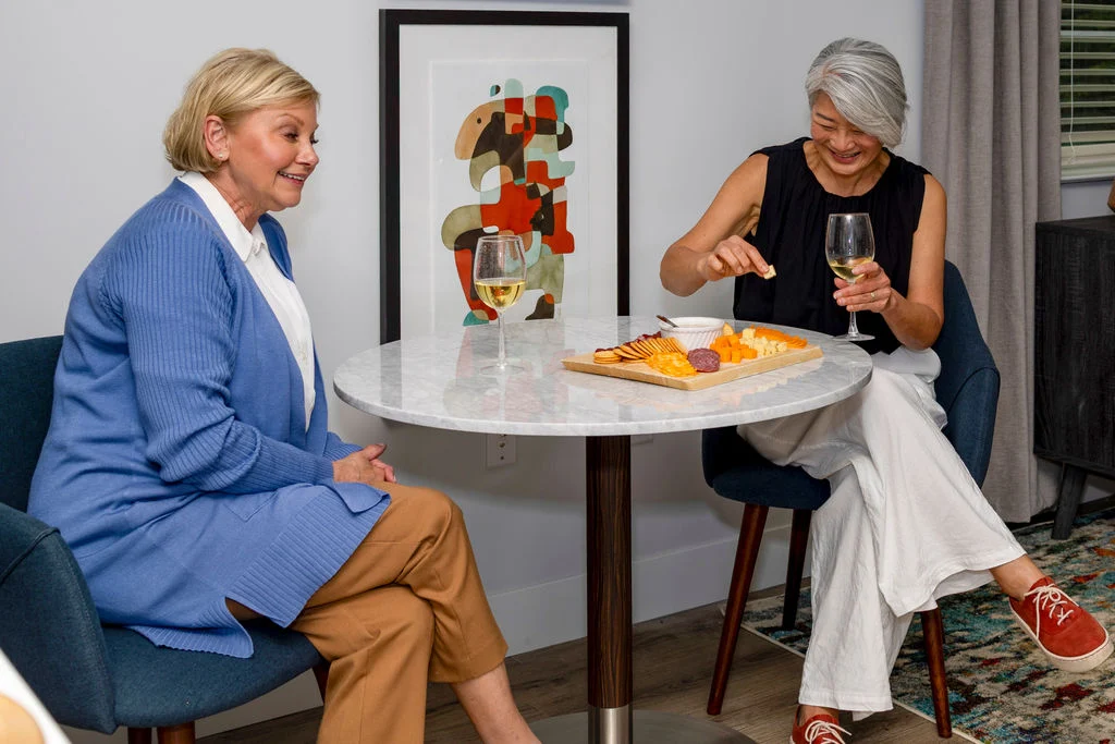 Two women sit at a small round table with wine and a cheese platter, smiling and chatting in a cozy, modern room at Verena at Hilliard, with abstract art on the wall behind them.