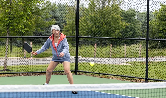 An older woman wearing a hoodie plays pickleball on an outdoor court, preparing to hit a yellow ball with her paddle. Trees and a fence are visible in the background.