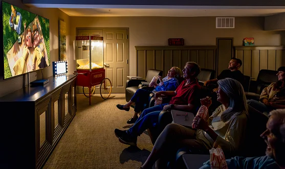 A group of six people sit in a cozy home theater watching a movie on a large screen, eating popcorn from bags, with a popcorn machine in the background and soft, dim lighting.