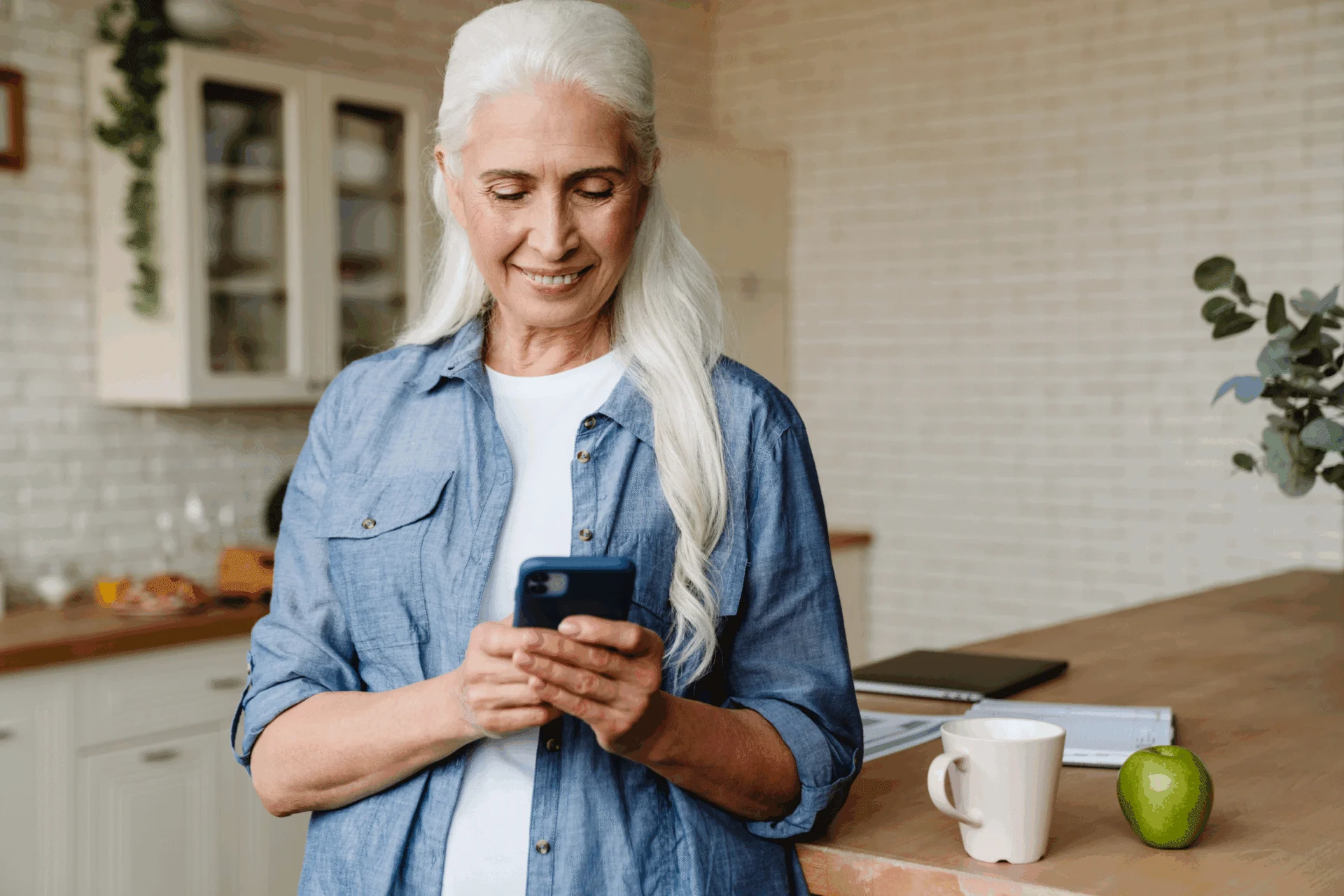 Senior woman with long gray hair looking at her smartphone in a bright kitchen
