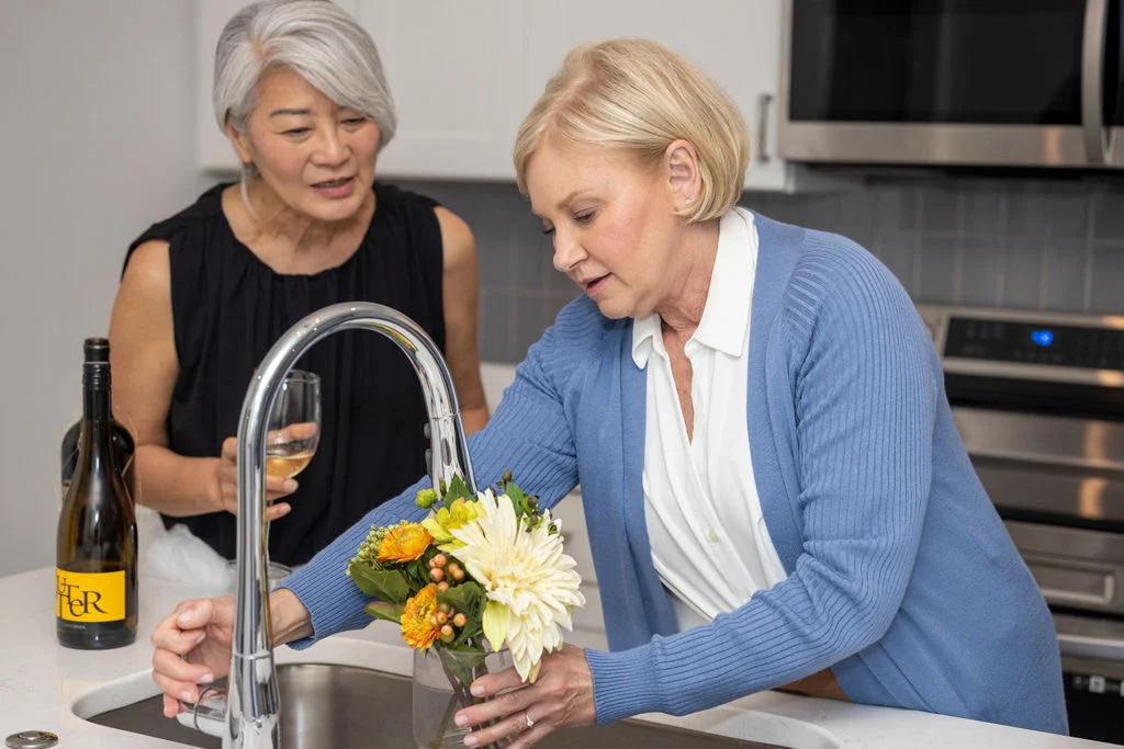 Two senior women arranging yellow flowers at a kitchen sink